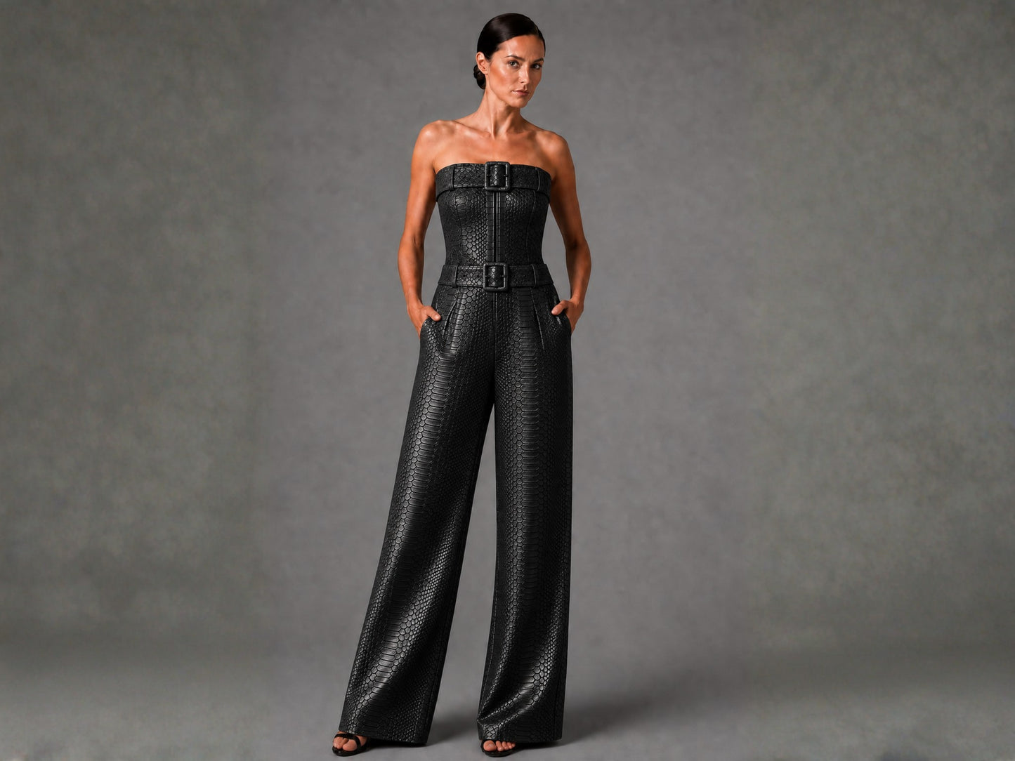 A woman modeling a black strapless wide-leg jumpsuit with a textured snakeskin pattern and double belt buckles at the waist, set against a neutral gray studio background.