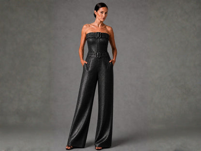 A woman modeling a black strapless wide-leg jumpsuit with a textured snakeskin pattern and double belt buckles at the waist, set against a neutral gray studio background.