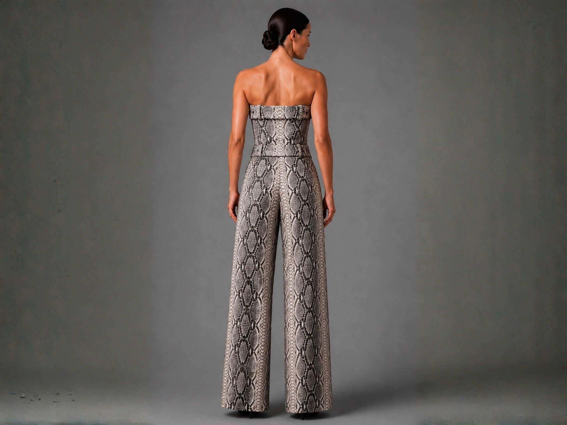 A woman shown from the back wearing a strapless wide-leg jumpsuit with a snakeskin pattern in beige, gray, and black, set against a solid gray studio background.