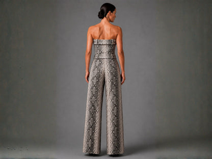 A woman shown from the back wearing a strapless wide-leg jumpsuit with a snakeskin pattern in beige, gray, and black, set against a solid gray studio background.
