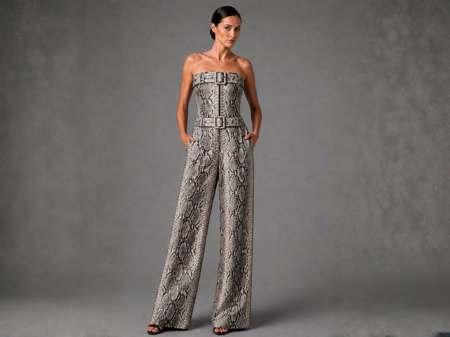 A woman models a strapless wide-leg jumpsuit with a snakeskin pattern and matching belt, standing against a textured gray studio backdrop.