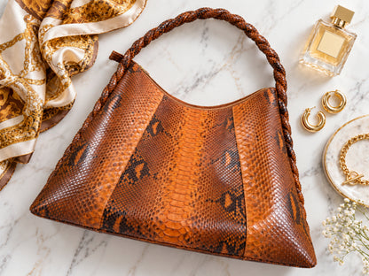 A brown and orange snakeskin-pattern shoulder bag with braided handles, styled on a white marble surface with a gold-and-white scarf, perfume bottle, earrings, and jewelry nearby.