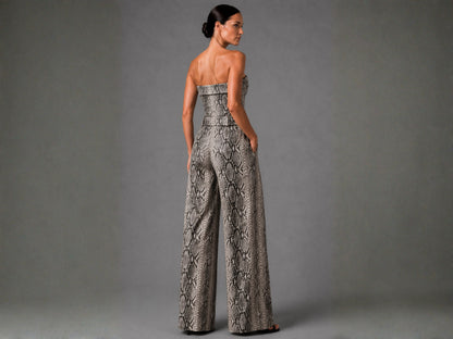 A woman stands with her back to the camera wearing a strapless wide-leg jumpsuit with a gray and beige snakeskin pattern, set against a solid gray studio background.
