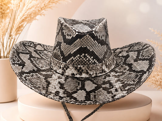 A wide-brimmed cowboy hat with a black and gray python-patterned design, displayed on a round beige pedestal with dried grass in the background.