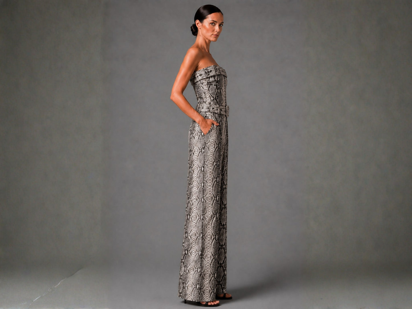 A woman modeling a strapless, floor-length gown with a gray and black snakeskin pattern, standing against a neutral gray studio backdrop.