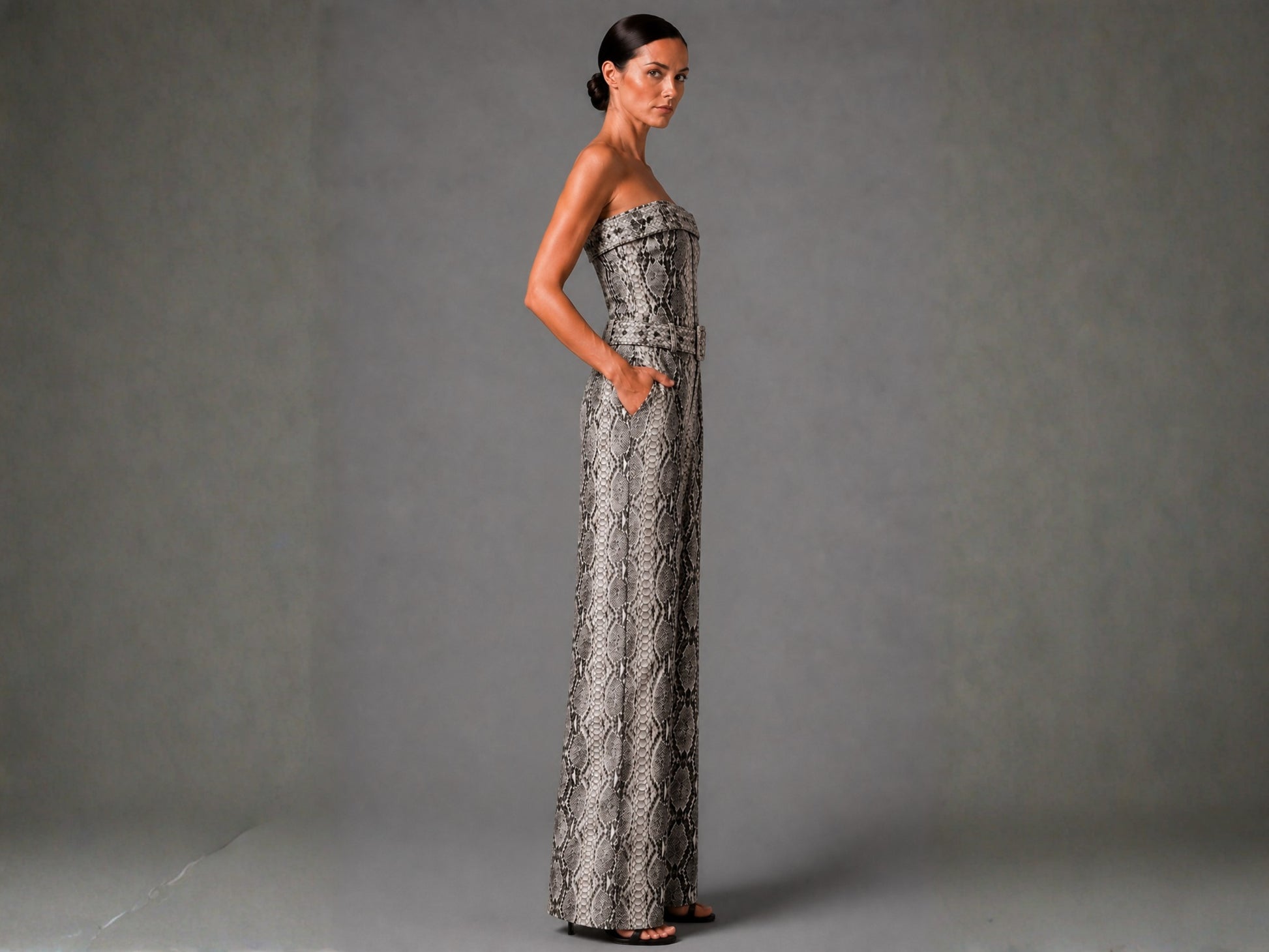 A woman modeling a strapless, floor-length gown with a gray and black snakeskin pattern, standing against a neutral gray studio backdrop.