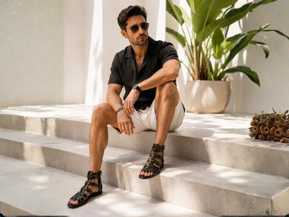 A man wearing black strappy gladiator sandals, a black short-sleeve button-up shirt, and off-white shorts sits on light stone steps in a sunlit outdoor setting with a potted plant in the background.