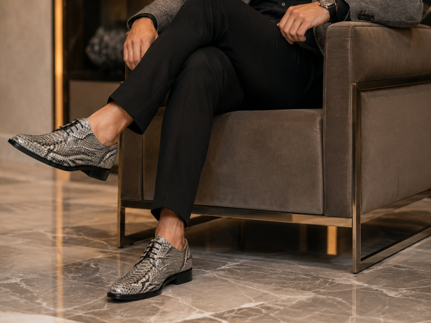 A man sitting in a modern brown armchair wearing black trousers and textured snakeskin-patterned dress shoes on a polished marble floor.
