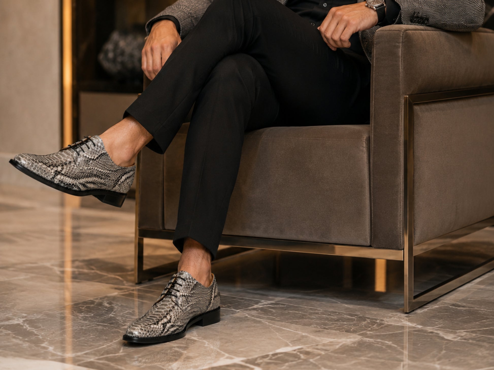 A man sitting in a modern brown armchair wearing black trousers and textured snakeskin-patterned dress shoes on a polished marble floor.