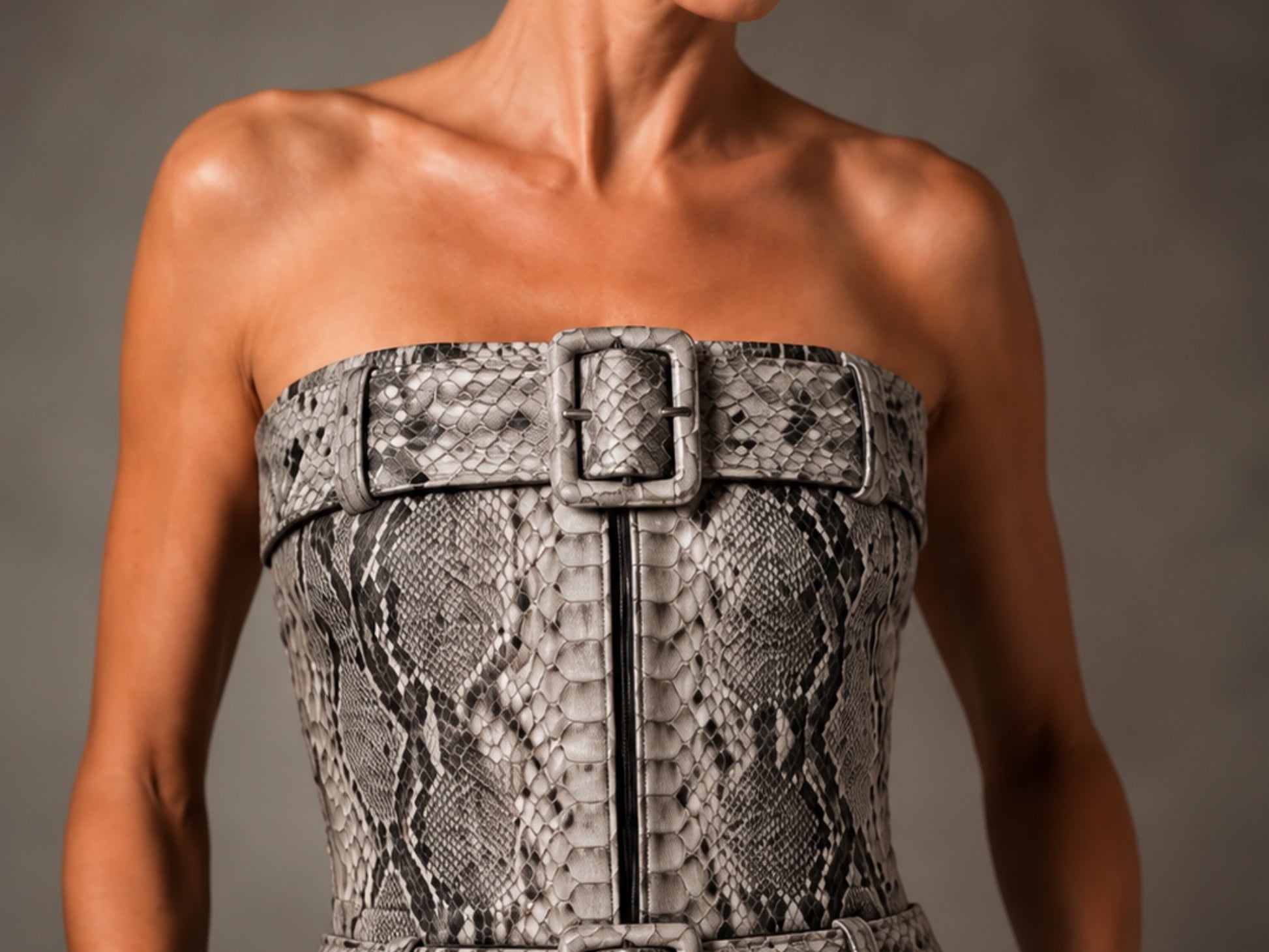 A close-up of a model wearing a strapless snakeskin-patterned corset-style top with a wide matching belt and large front buckle, against a neutral gray studio background.