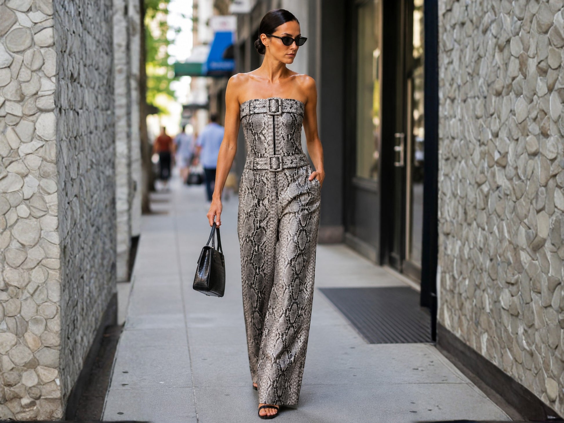 A woman walks down a city sidewalk wearing a strapless snakeskin-pattern jumpsuit in gray and beige tones, holding a black handbag.