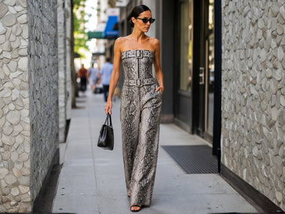 A woman walks down a city sidewalk wearing a strapless snakeskin-pattern jumpsuit in gray and beige tones, holding a black handbag.