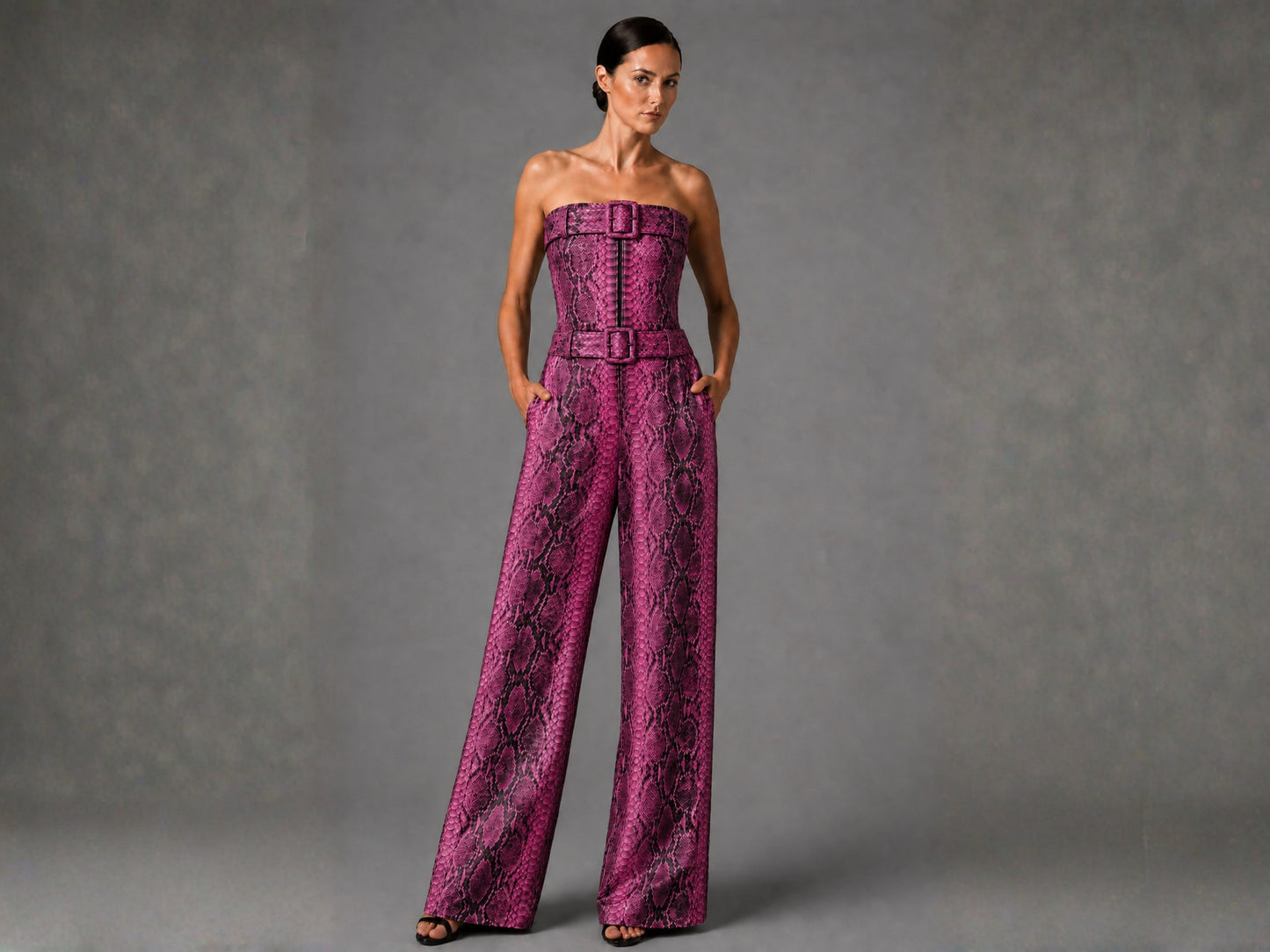 A woman modeling a strapless wide-leg jumpsuit with a bold pink and black snakeskin pattern, standing against a textured gray studio background.