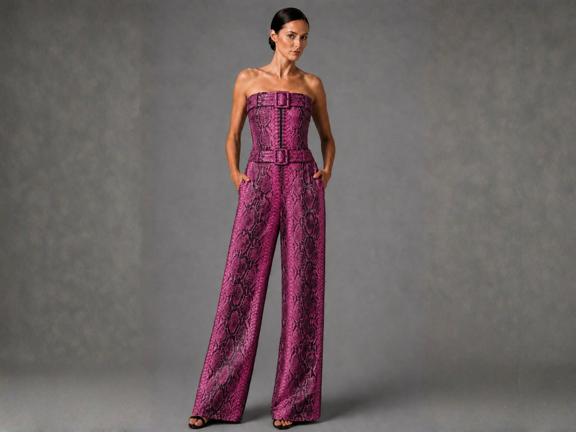 A woman modeling a strapless wide-leg jumpsuit with a bold pink and black snakeskin pattern, standing against a textured gray studio background.