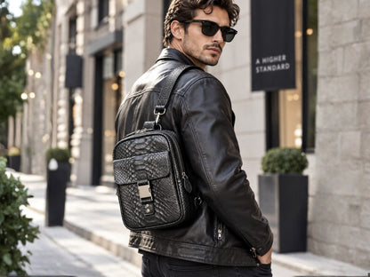 A man wearing a black leather jacket and sunglasses carries a black snakeskin-leather crossbody bag with silver hardware on a city street.