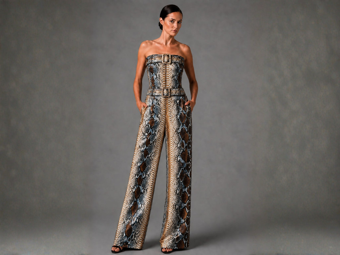 A woman modeling a strapless wide-leg jumpsuit with a snakeskin pattern in brown, beige, and gray tones, featuring a structured waist with belt-like detailing. She is standing against a textured gray studio backdrop.