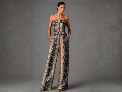 A woman modeling a strapless wide-leg jumpsuit with a snakeskin pattern in brown, beige, and gray tones, featuring a structured waist with belt-like detailing. She is standing against a textured gray studio backdrop.