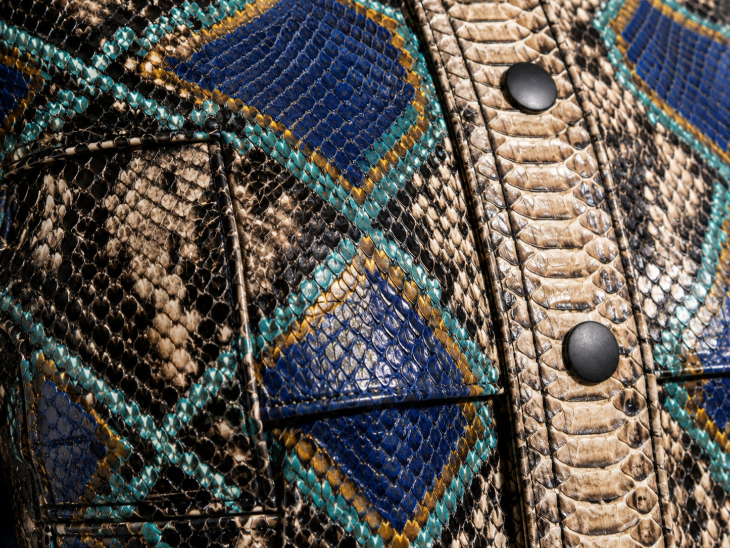 Close-up of a textured leather jacket featuring a geometric diamond pattern with blue, teal, and tan snakeskin print, accented by black snap buttons.