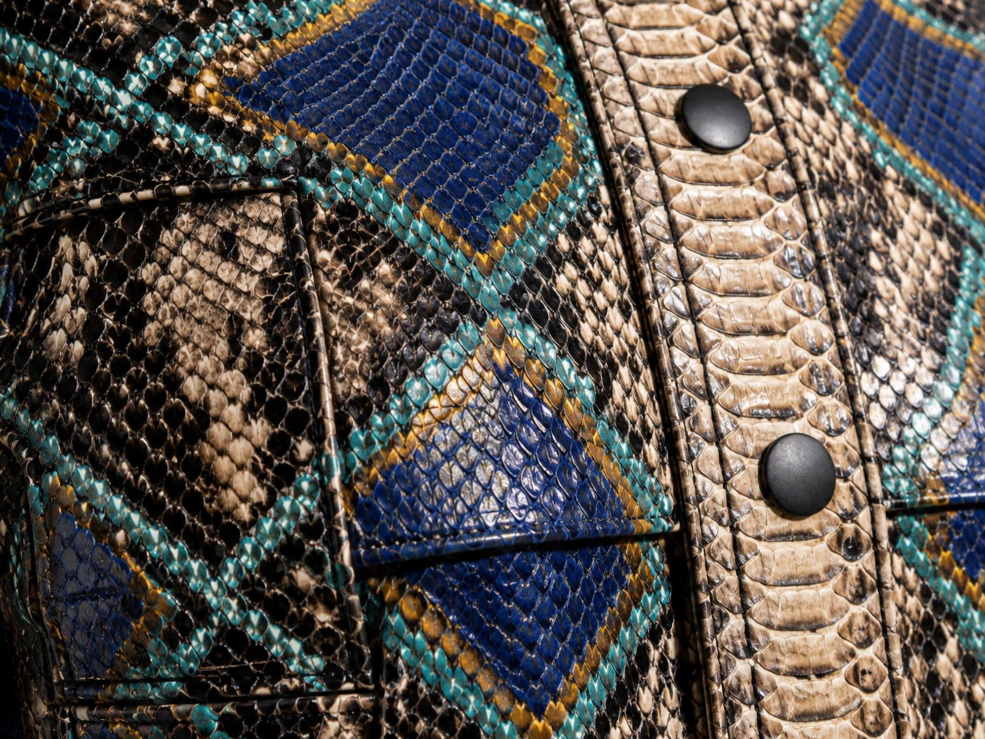 Close-up of a textured leather jacket featuring a geometric diamond pattern with blue, teal, and tan snakeskin print, accented by black snap buttons.