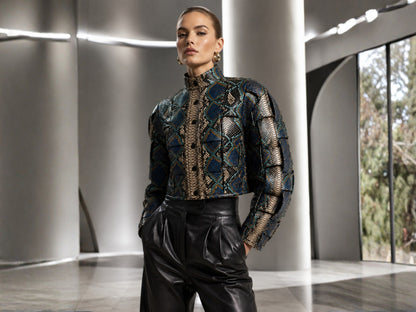A model wearing a high-neck, long-sleeve cropped jacket with a complex geometric pattern in blue, gold, and black, paired with black leather pants, standing in a modern architectural hallway.