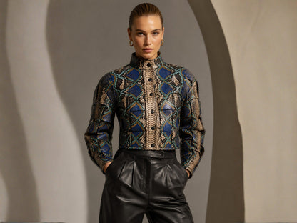 A model wearing a high-neck, long-sleeve button-front jacket with a blue, black, and beige geometric pattern, paired with black leather pants, set against a gray studio backdrop with dramatic shadows.